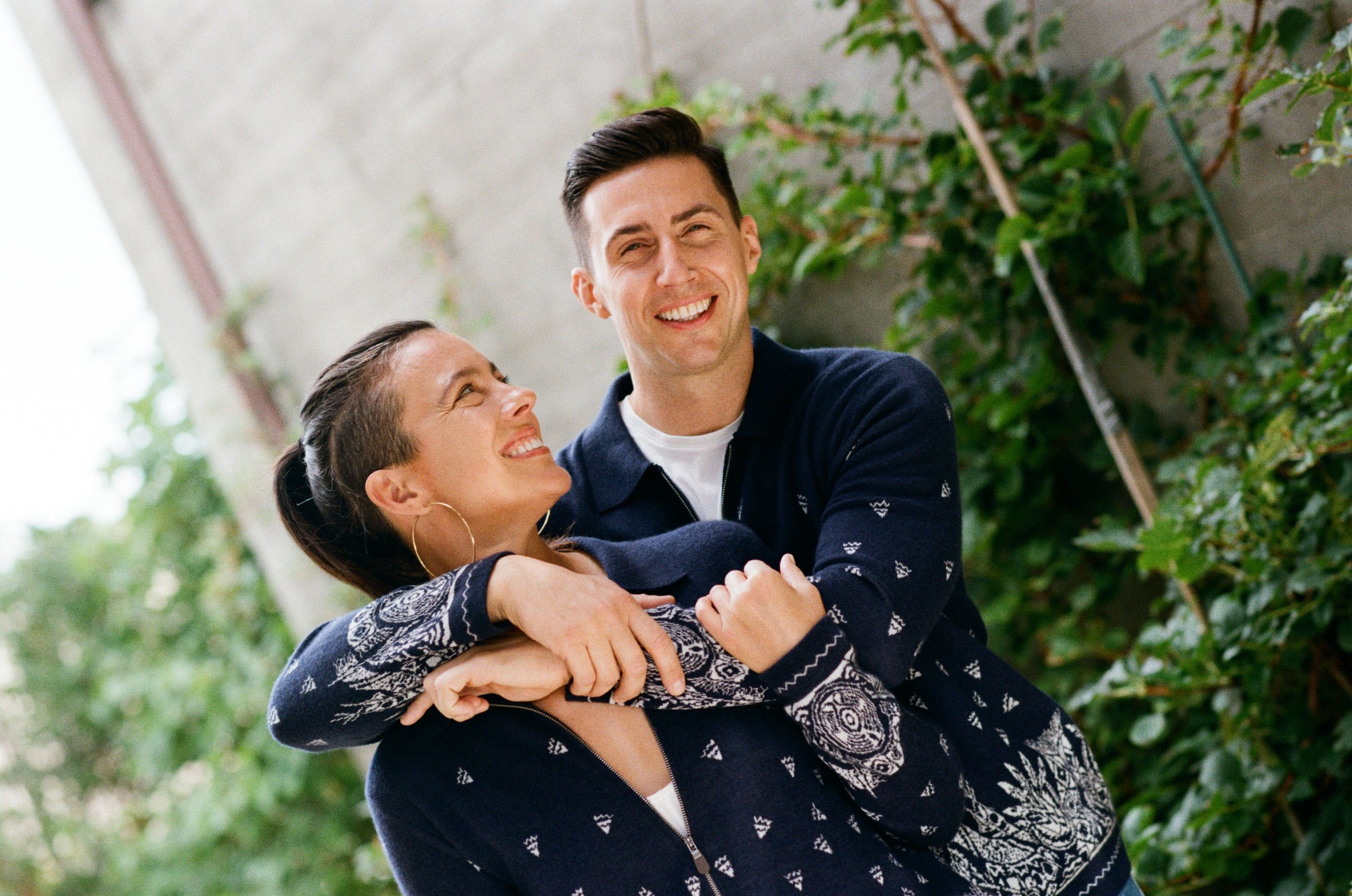 Two people wearing cashmere cardigans with bandana patterns, embracing outdoors with greenery in the background