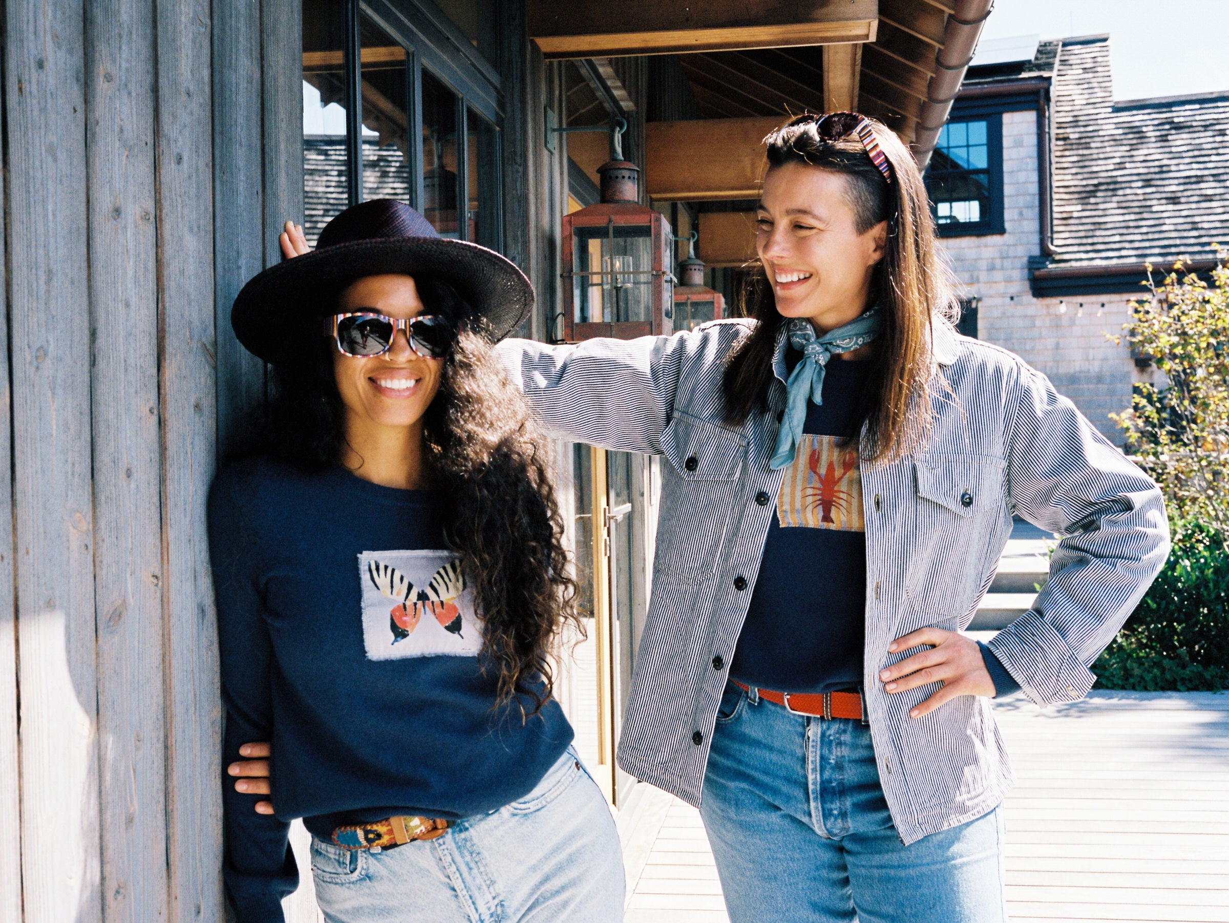 Two people standing outside a building, one wearing a hat, cashmere sweater, and sunglasses, the other with a striped jacket.