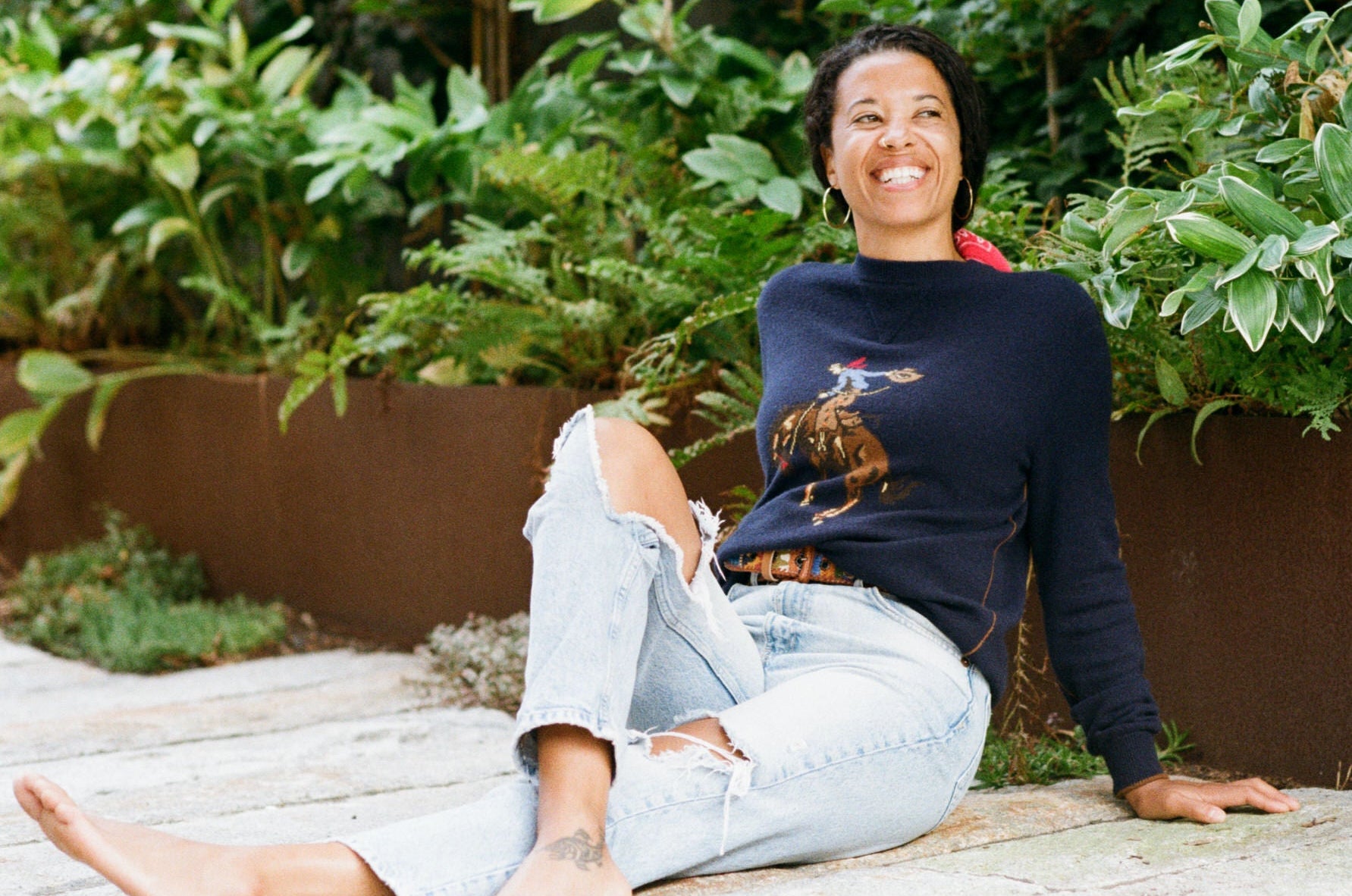 Woman wearing a cashmere sweater with a cowboy pattern sitting outdoors with plants in the background