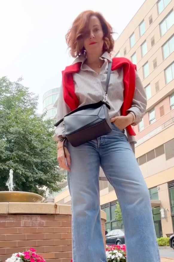 Woman with red hair wearing a red cashmere Daniel Hammond sweater, white oxford shirt, and blue jeans with a black handbag outdoors in the city.